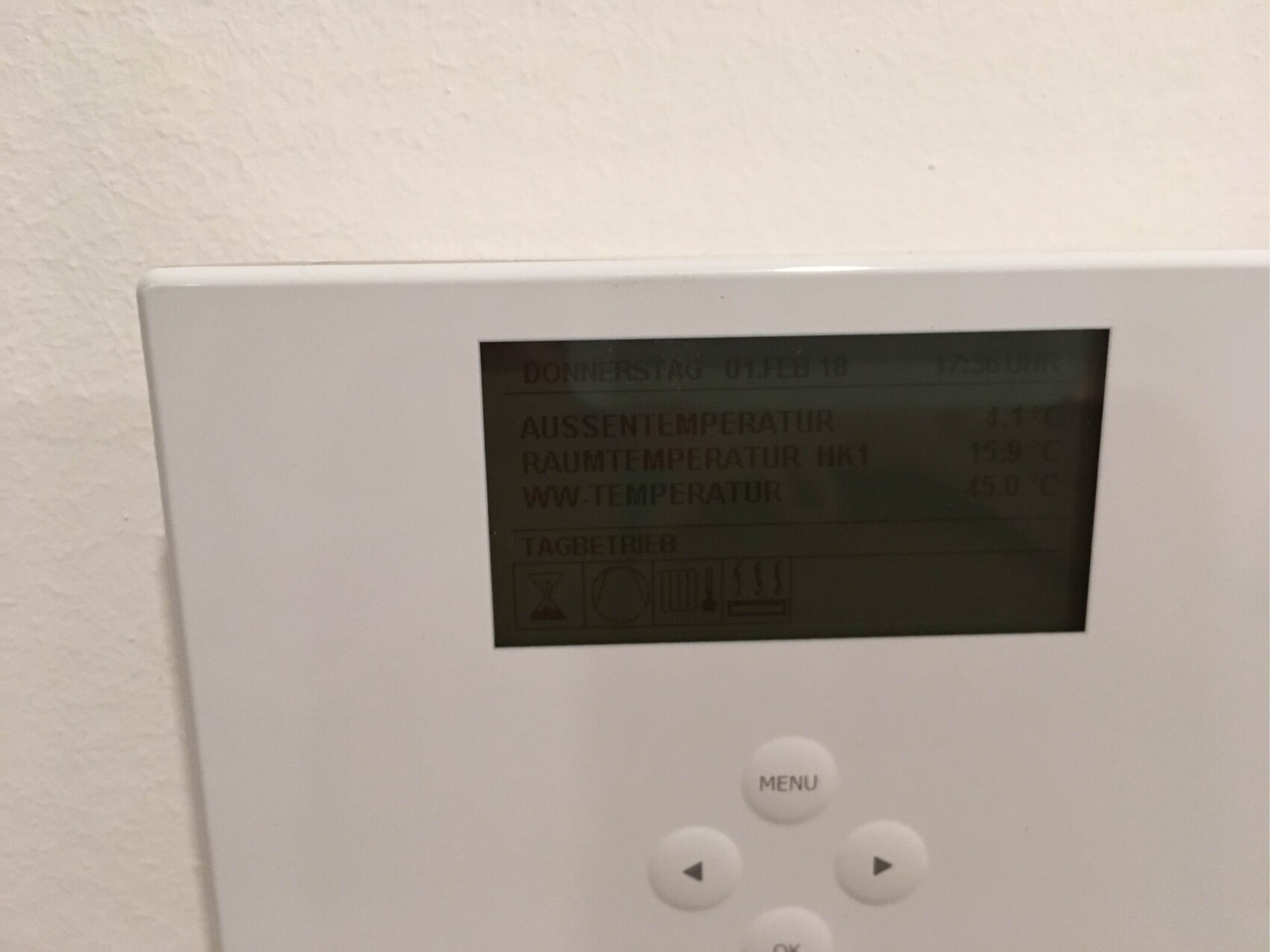 Weißes Wandthermostat mit digitaler Anzeige und Menüknopf an der Wand.
