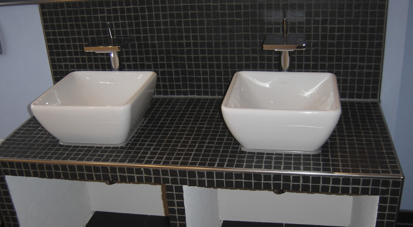 Zwei weiße quadratische Waschbecken auf einer schwarzen Kacheltischplatte mit Chrom-Armaturen.
