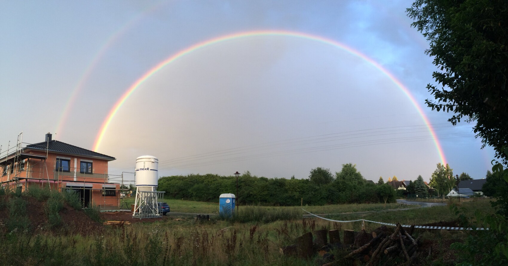 Zweistöckiges Haus im Rohbau mit Gerüst, Baucontainer und Doppelregenbogen am Himmel