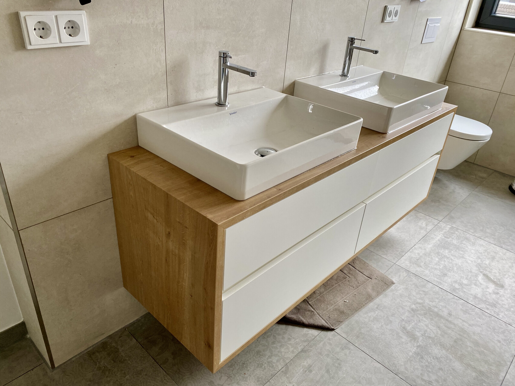Zwei weiße Keramik-Waschbecken auf Holz-Unterschrank in modernem Badezimmer, Toilette rechts.