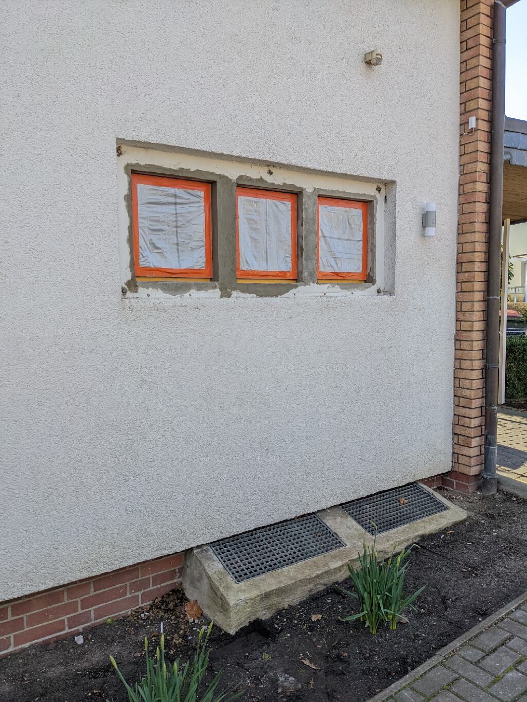 Weiße Außenwand mit drei orange gerahmten Fenstern hinter Folie; unten Lüftungsgitter.