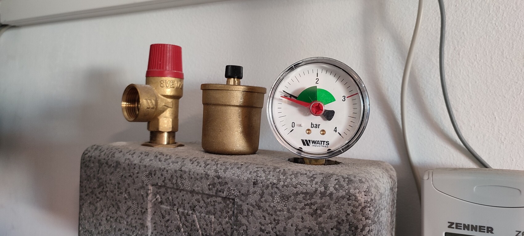 Druckmessgerät (bar) neben Messingventil mit rotem Deckel und Zylinder auf grauer Platte.