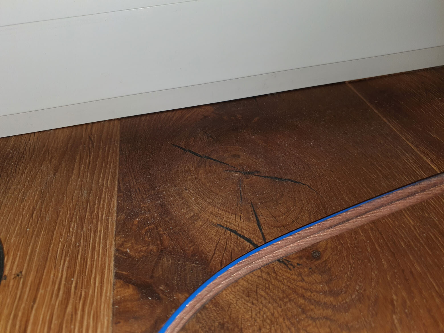Holzboden mit dunklen Kratzern und Rissen, weiße Sockelleiste; blaues Kabel liegt darüber.