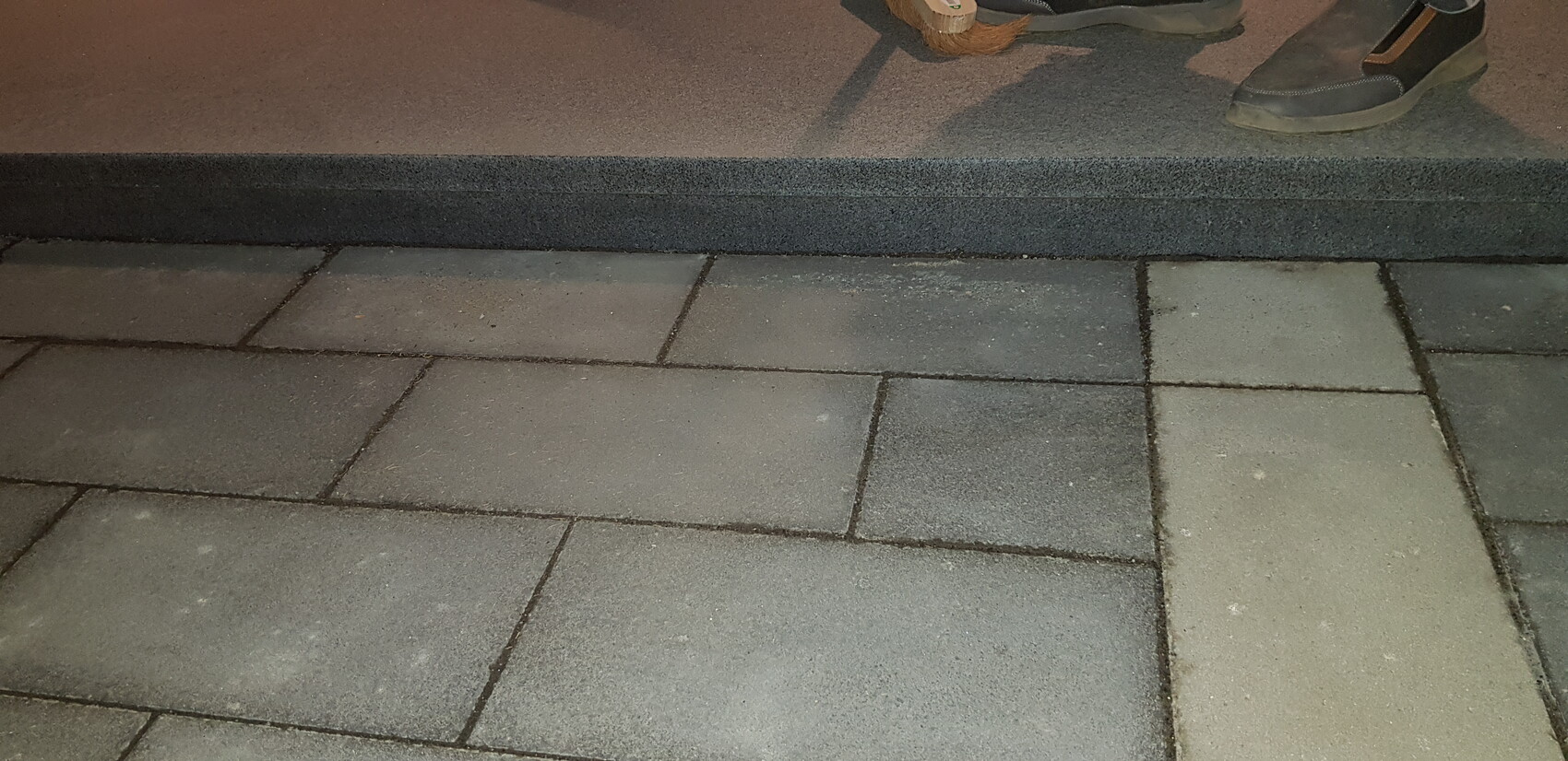 Grauer Pflasterboden mit dunkler Stufe; Schuh und Besen am oberen Rand sichtbar.