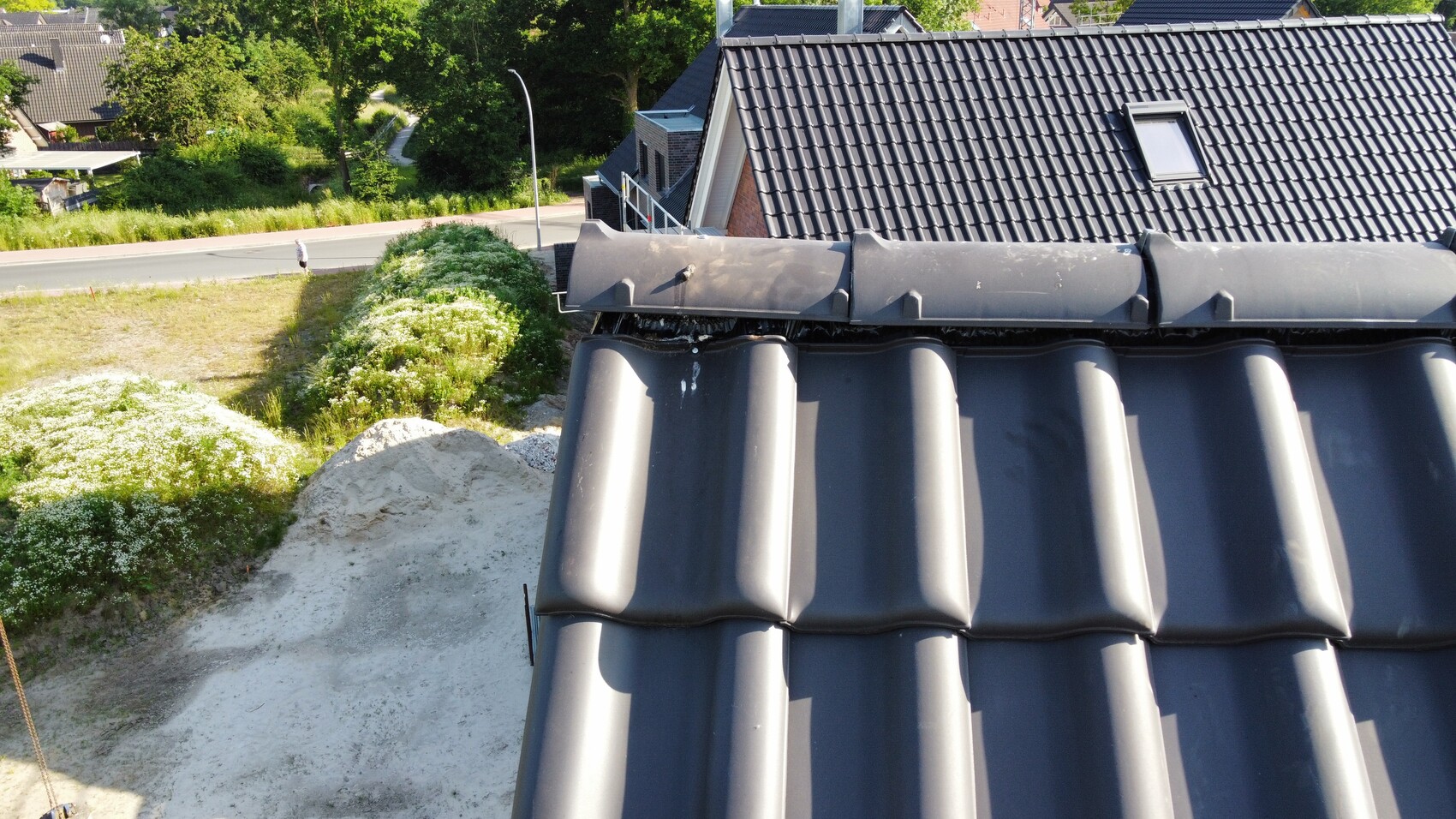 Draufsicht auf dunkle, wellige Dachziegel; Straße, Grünflächen, Sandhügel, kleine Person am Gehweg.