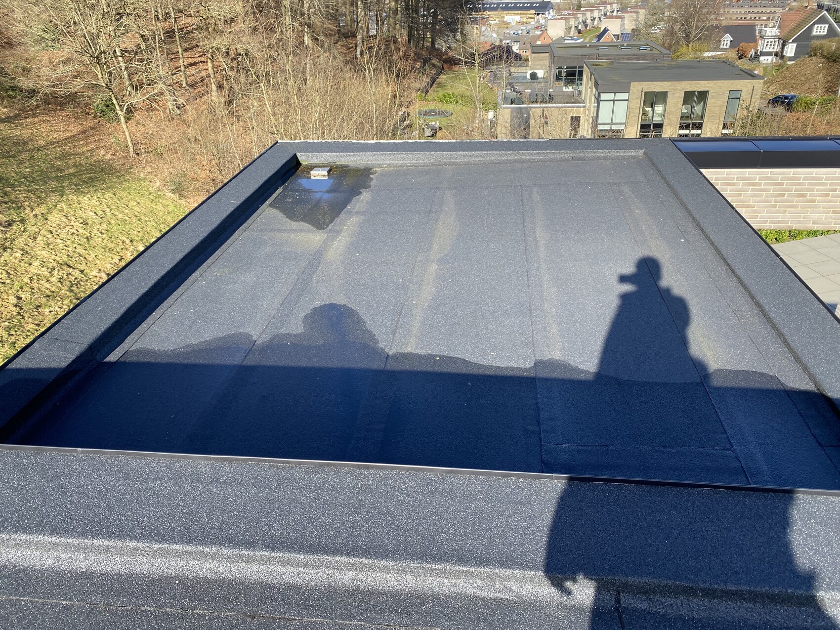 Draufsicht auf dunkles Flachdach, Schatten des Fotografen, städtische Häuser im Hintergrund.