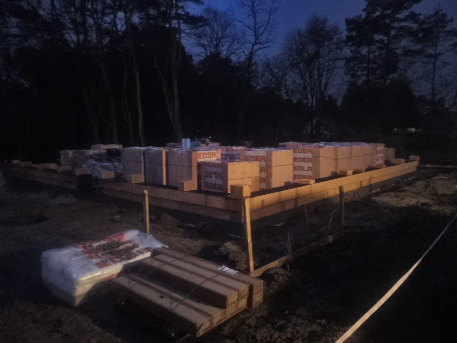 Baugrundstück mit gestapelten Steinen auf Holzfundament, Dämmerung, Wald im Hintergrund.