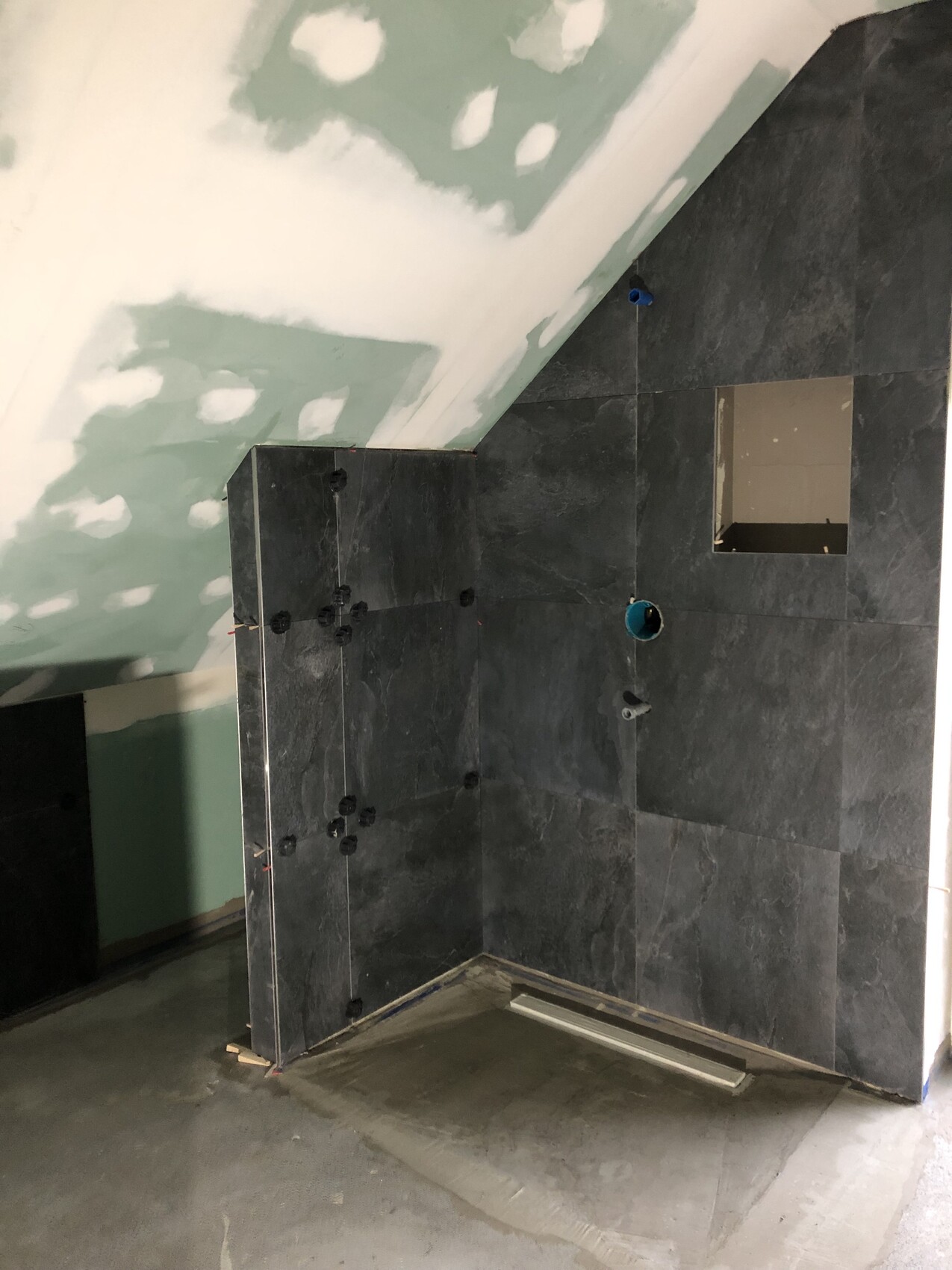 Unfertiger Badezimmerbereich mit dunklen Fliesen, schräger Decke und sichtbaren Rohren.