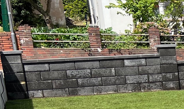 Grau-schwarze Steinmauer mit roten Ziegelpfeilern und Metallgeländer; Gras davor, Bäume dahinter.
