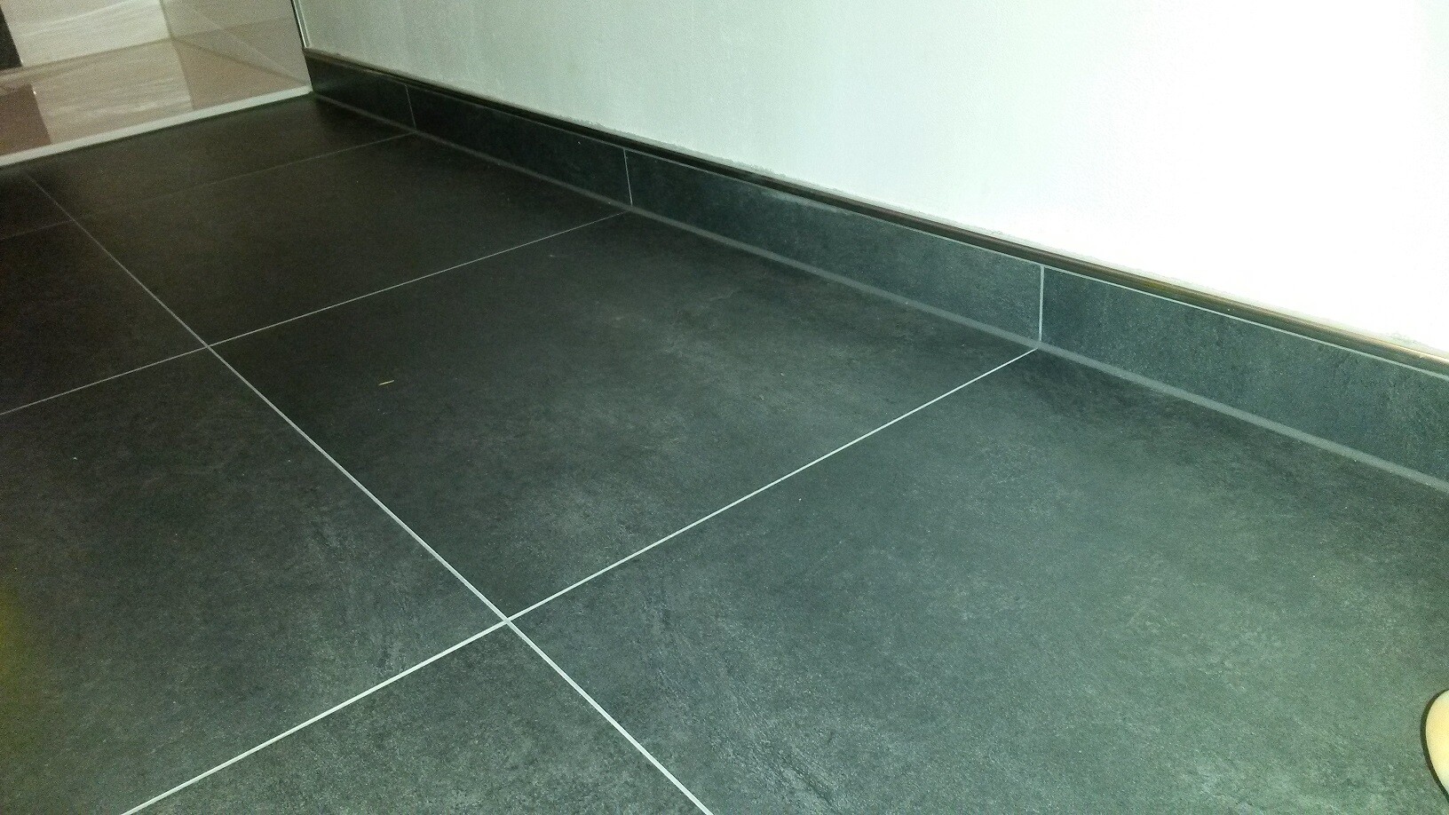 Dunkle quadratische Bodenfliesen mit hellen Fugen, weiße Wand und dunkler Sockelbereich.