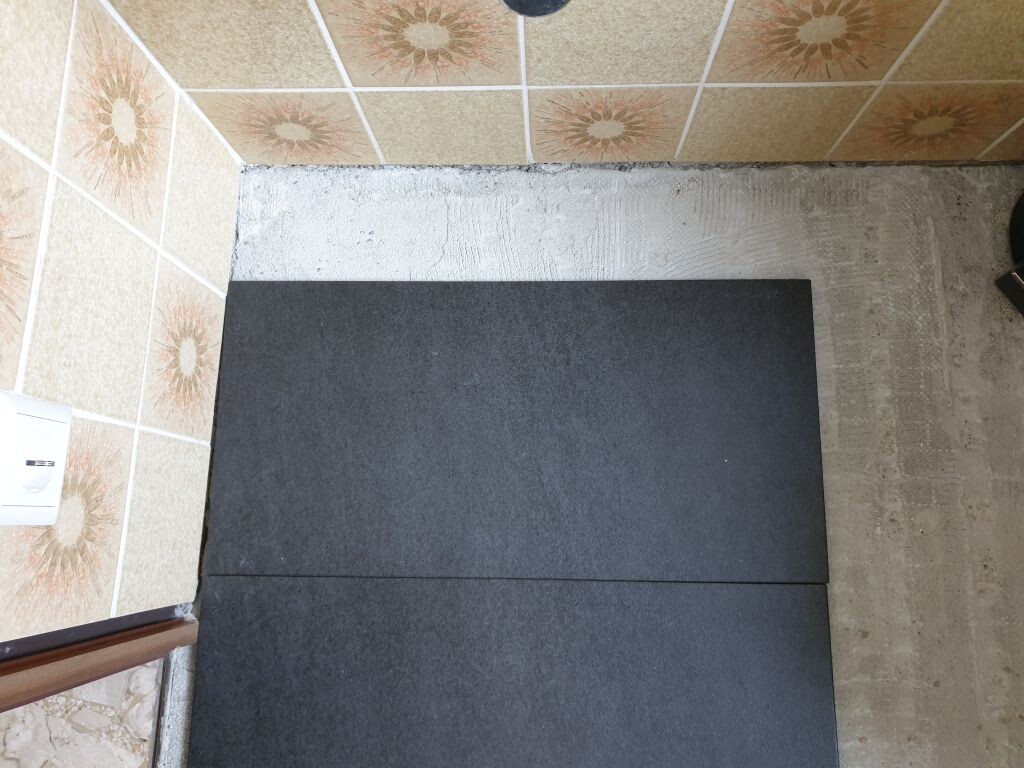 Zwei dunkelgraue Fliesen liegen auf Betonboden neben gemusterten Wandfliesen; Steckdose sichtbar.
