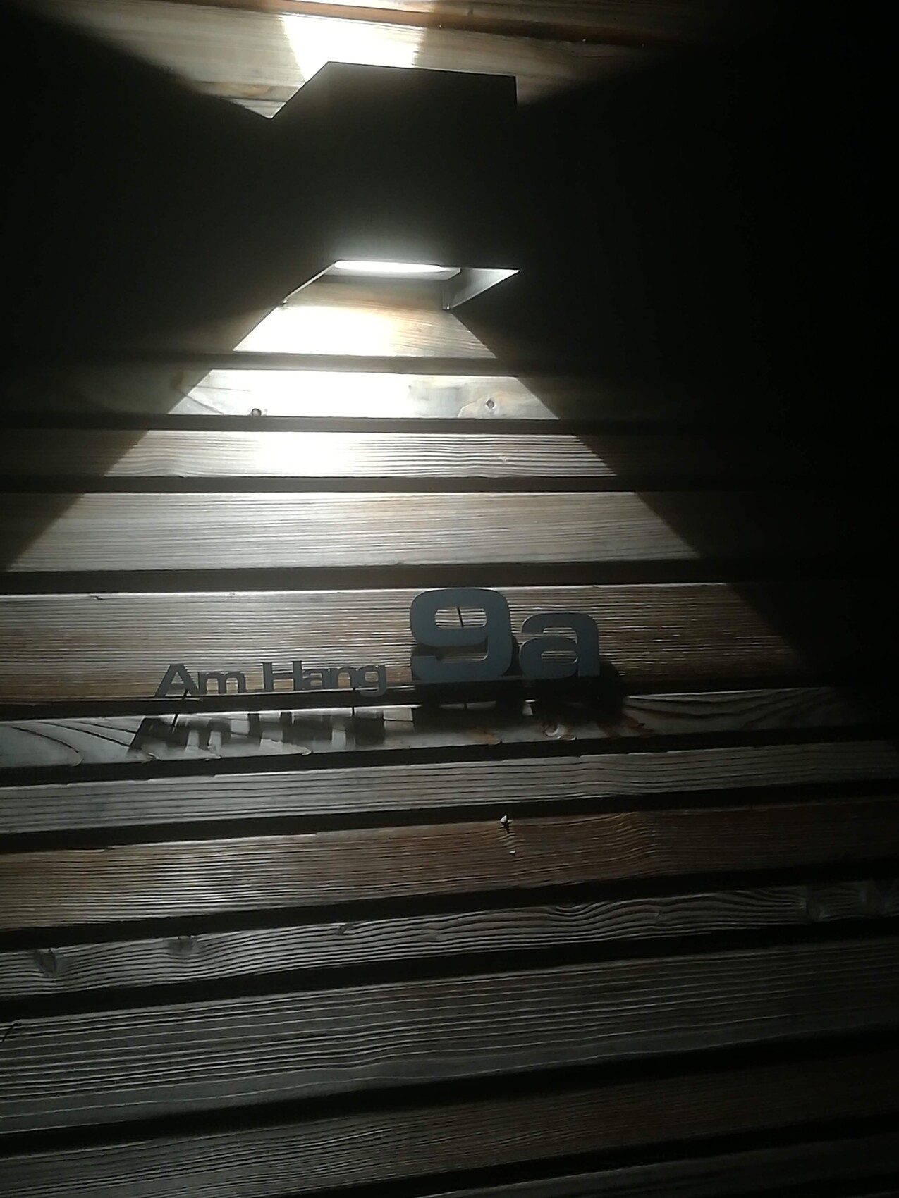 Holzverkleidete Wand mit schrägen Lichtstrahlen durch Öffnungen; Schild „Am Hang 9a“ sichtbar.