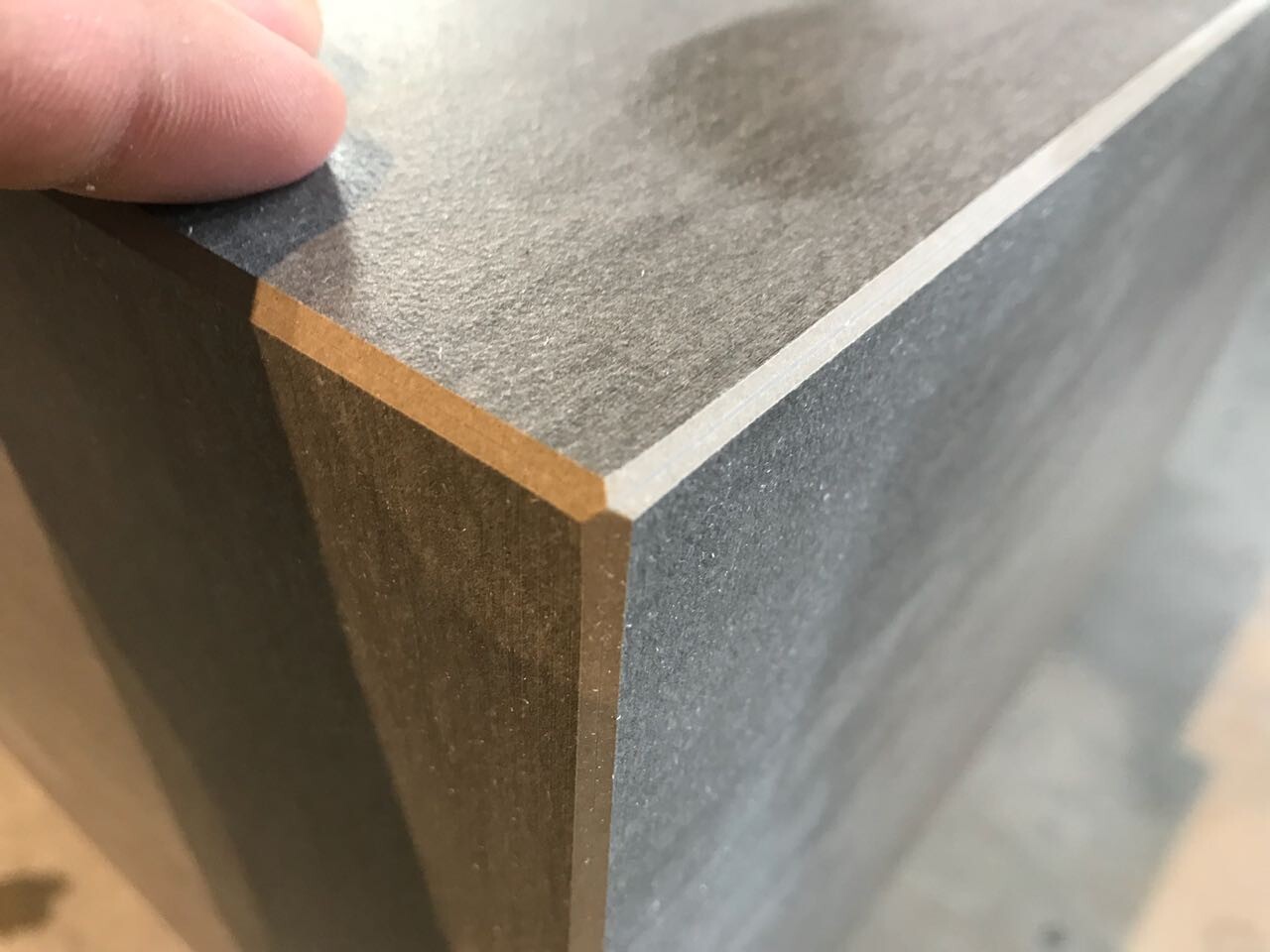 Nahaufnahme einer Holzplatte: graue Oberseite, braune Kante, Finger an der Ecke.