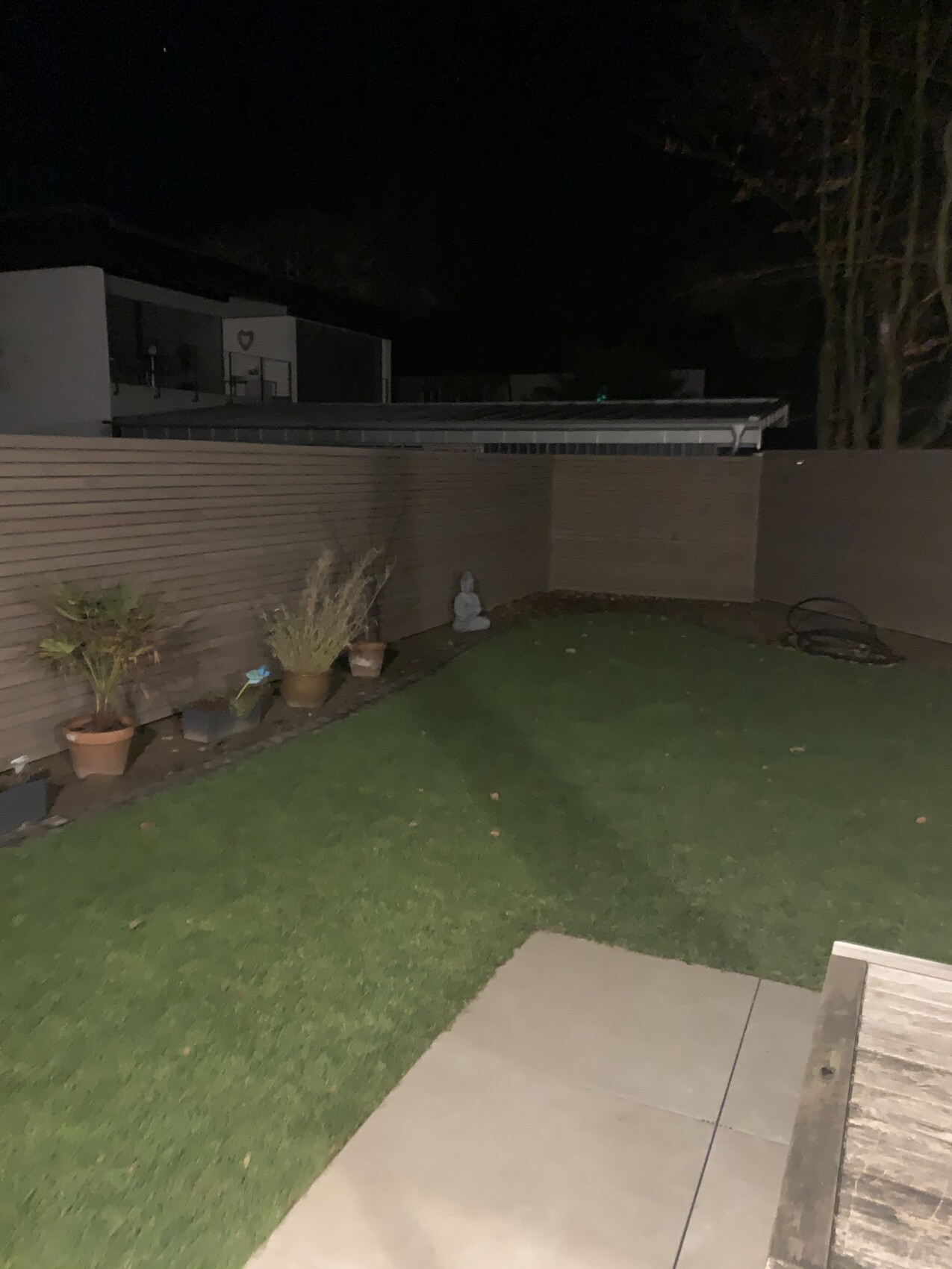 Garten bei Nacht: Grasfläche, Holzzaun, Topfpflanzen und eine Buddha-Statue am Rand.