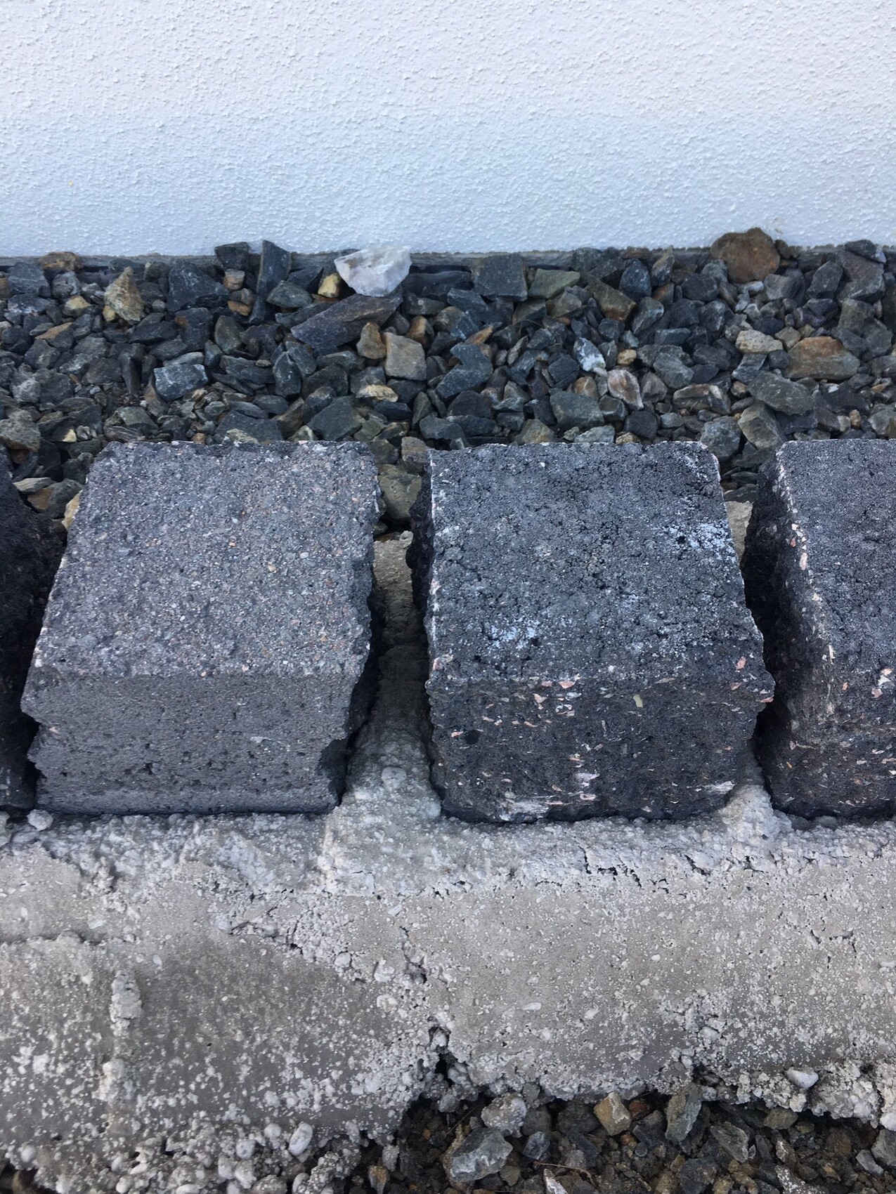 Große dunkle Pflastersteine werden als Terrassenboden neben Kies und einer weißen Wand verlegt.