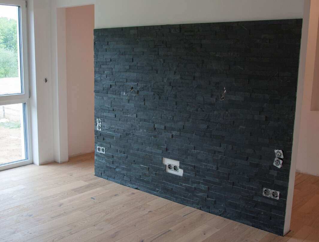 Eine dunkle Steinwand im Raum mit mehreren Steckdosen, Holzboden und großes Fenster links.