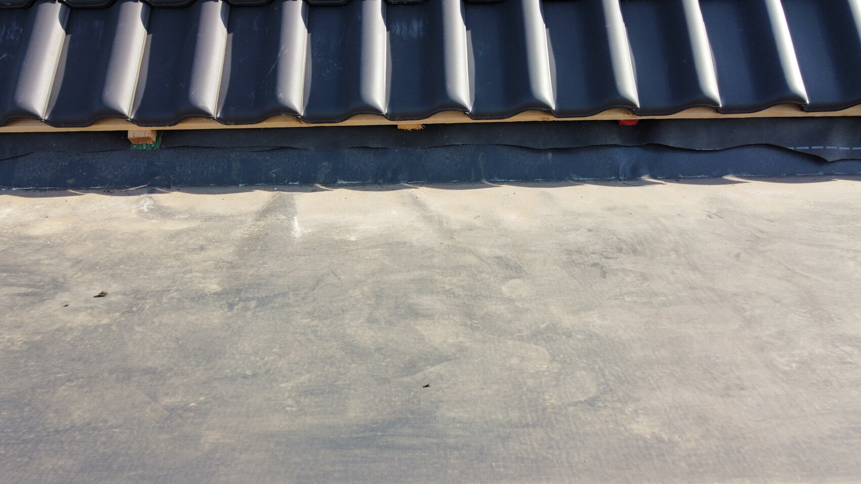 Schwarze, gewölbte Dachziegel am oberen Rand über dunkler Unterlage; darunter Betonfläche.