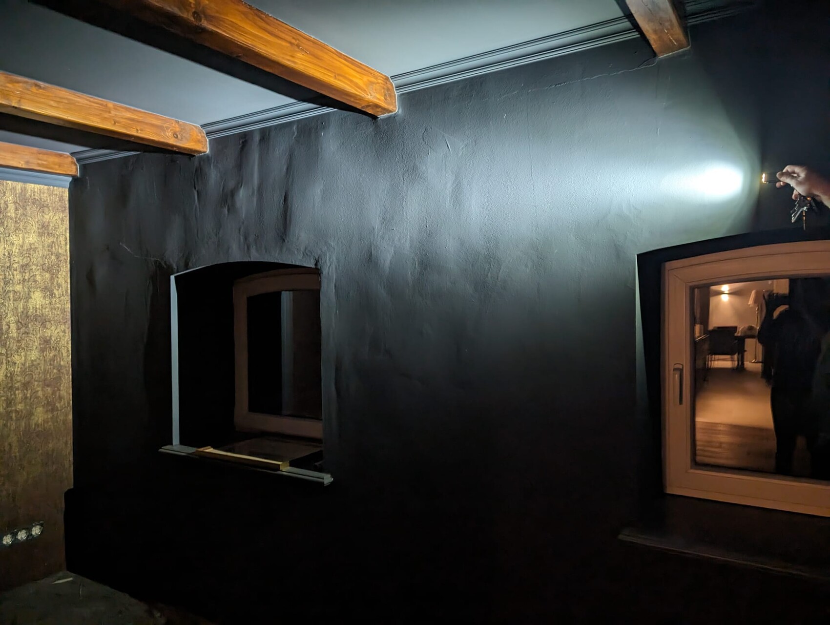 Dunkler Raum mit blauer Wand und Holzbalkendecke; rechts Lichtstrahl einer Taschenlampe.