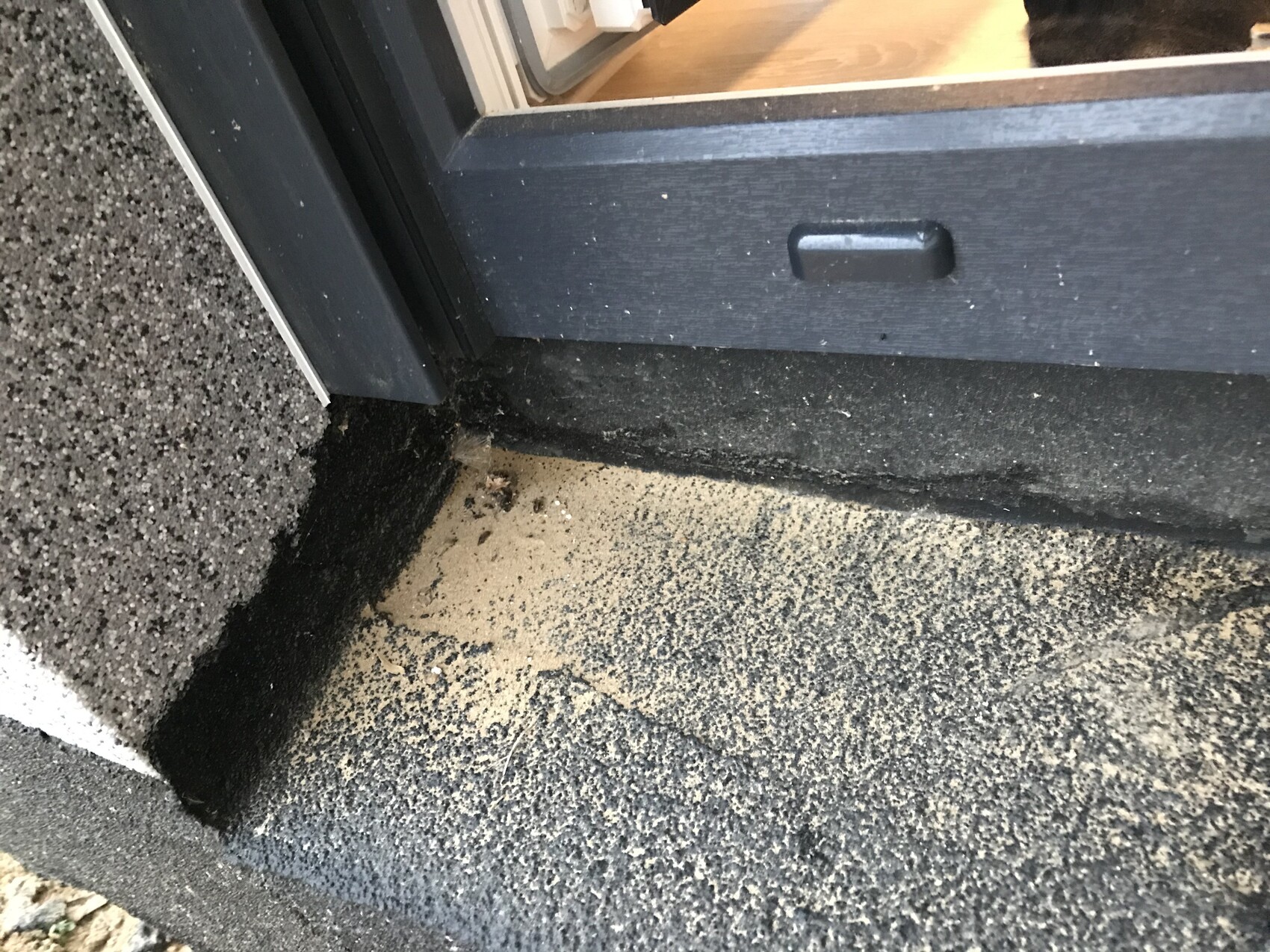 Außenbereich einer Türschwelle mit Sand auf dunkler Bodenplatte; Innen Holzboden sichtbar.