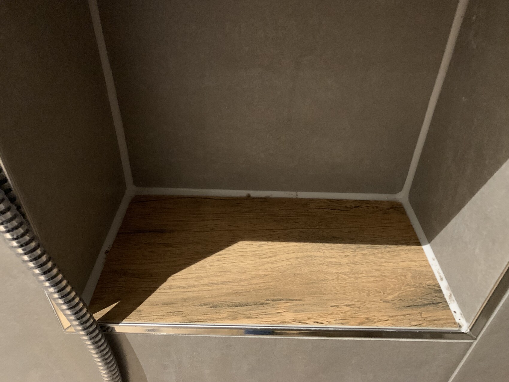 Ecke einer Dusche mit grauen Wänden, Holzoptik-Duschboden und flexibler Brauseschlauch
