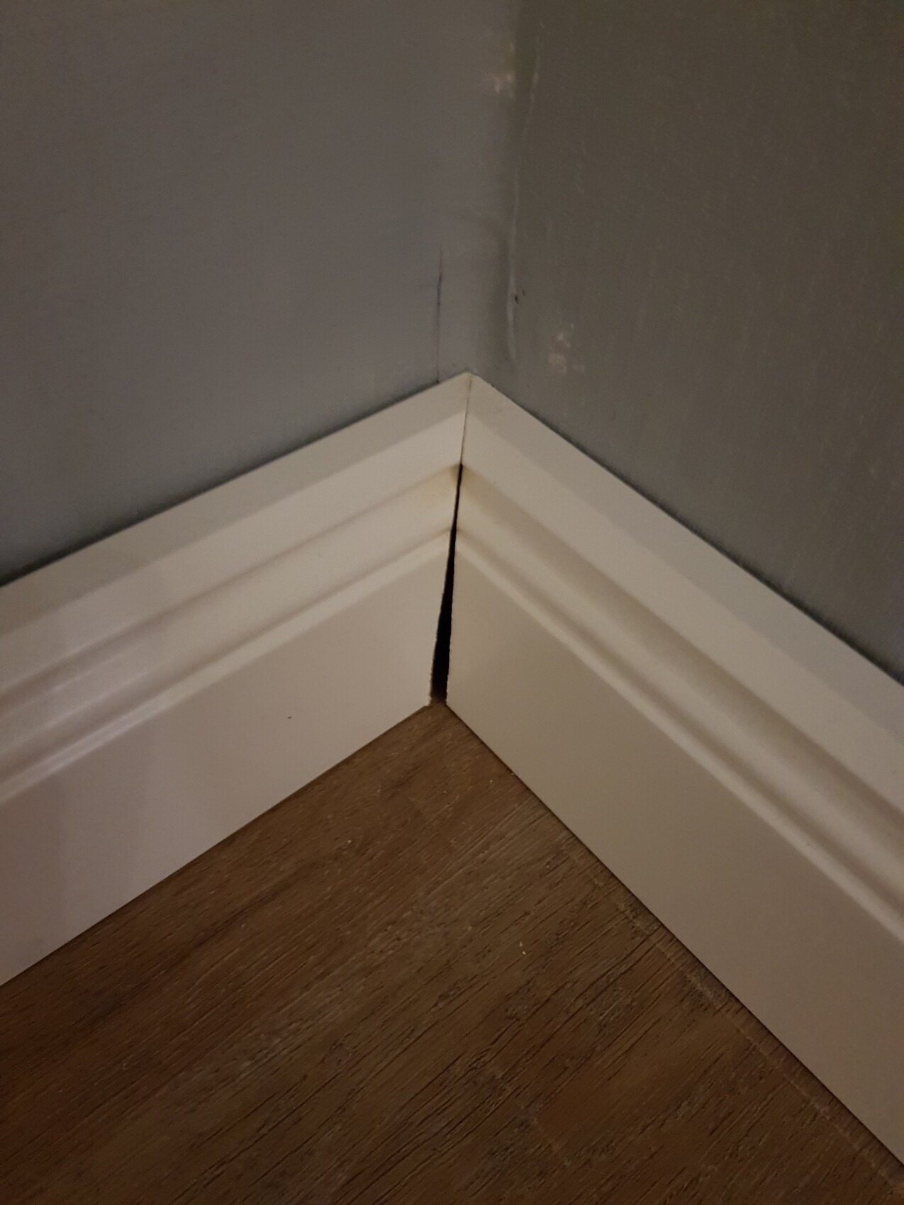 Eckbereich eines Zimmers: weiße Fußleiste mit Riss an der Eckverbindung, graue Wand, Holzboden.