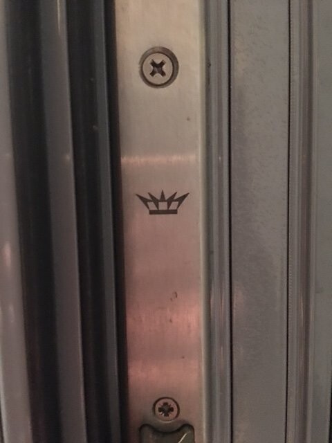 Nahaufnahme eines metallischen Türbeschlags mit Kronenemblem und Schrauben