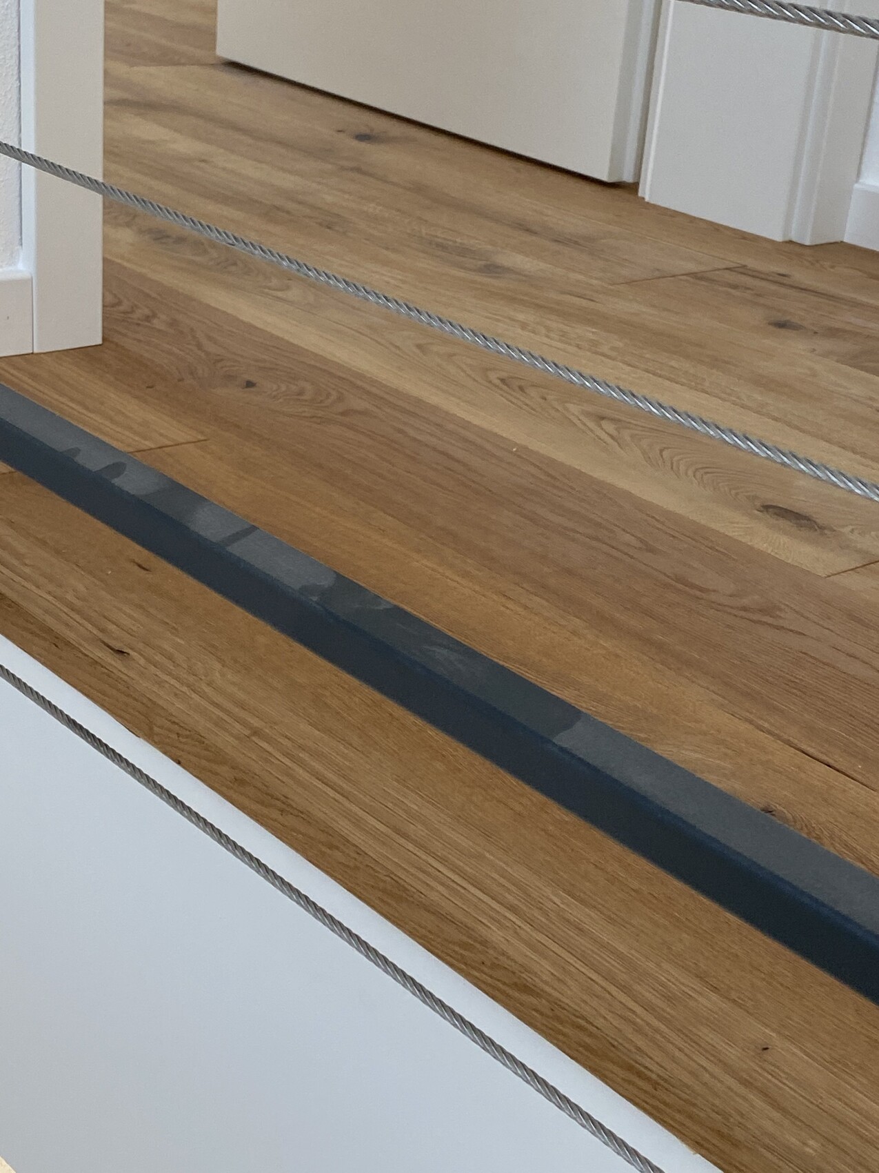 Holzboden mit zwei diagonalen Drahtseil-Geländern; weiße Türrahmen im Hintergrund.