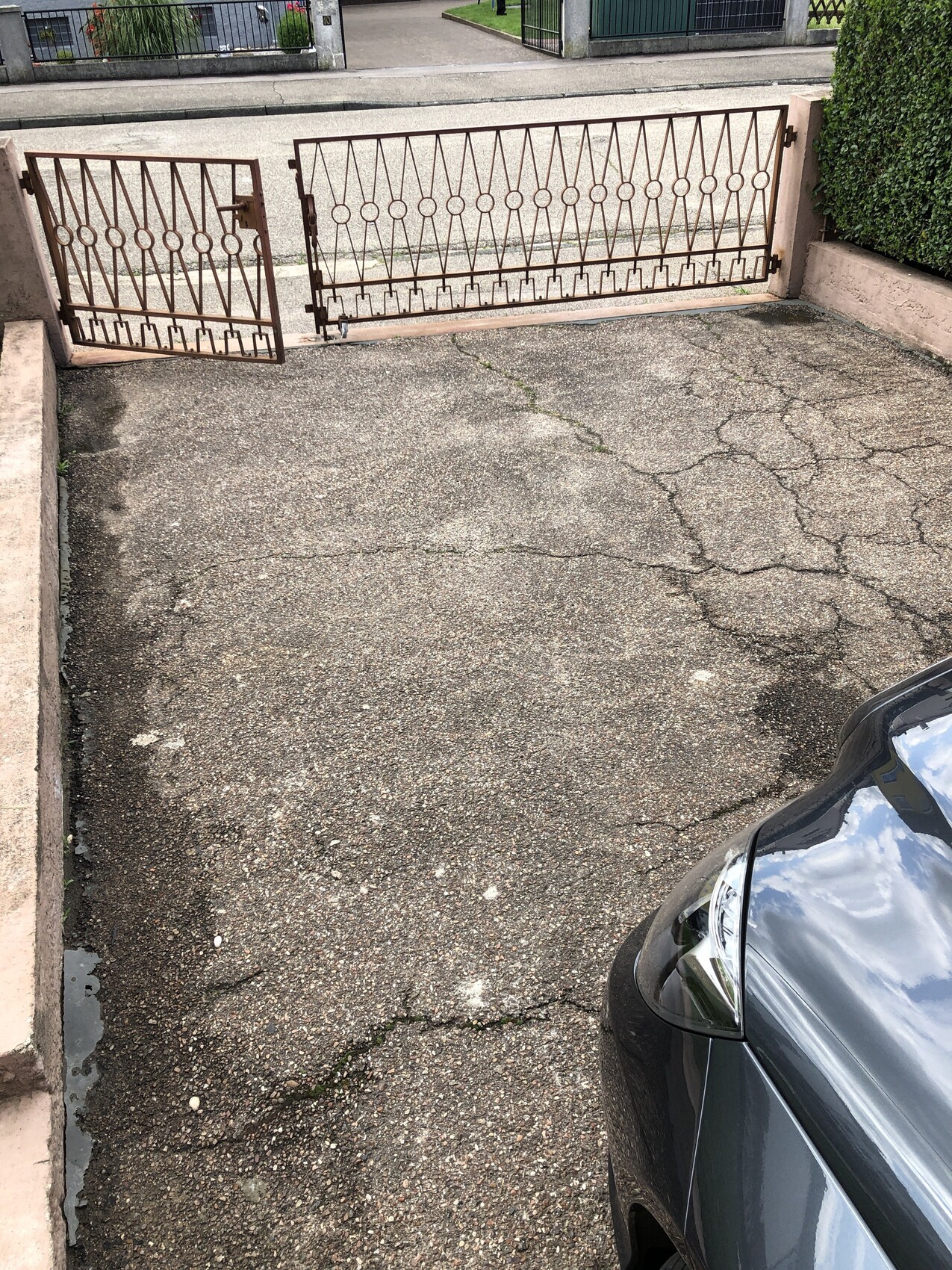 Geöffnete schmiedeeiserne Einfahrtstore, rissiger Asphalt, rechts vorne geparktes Auto.