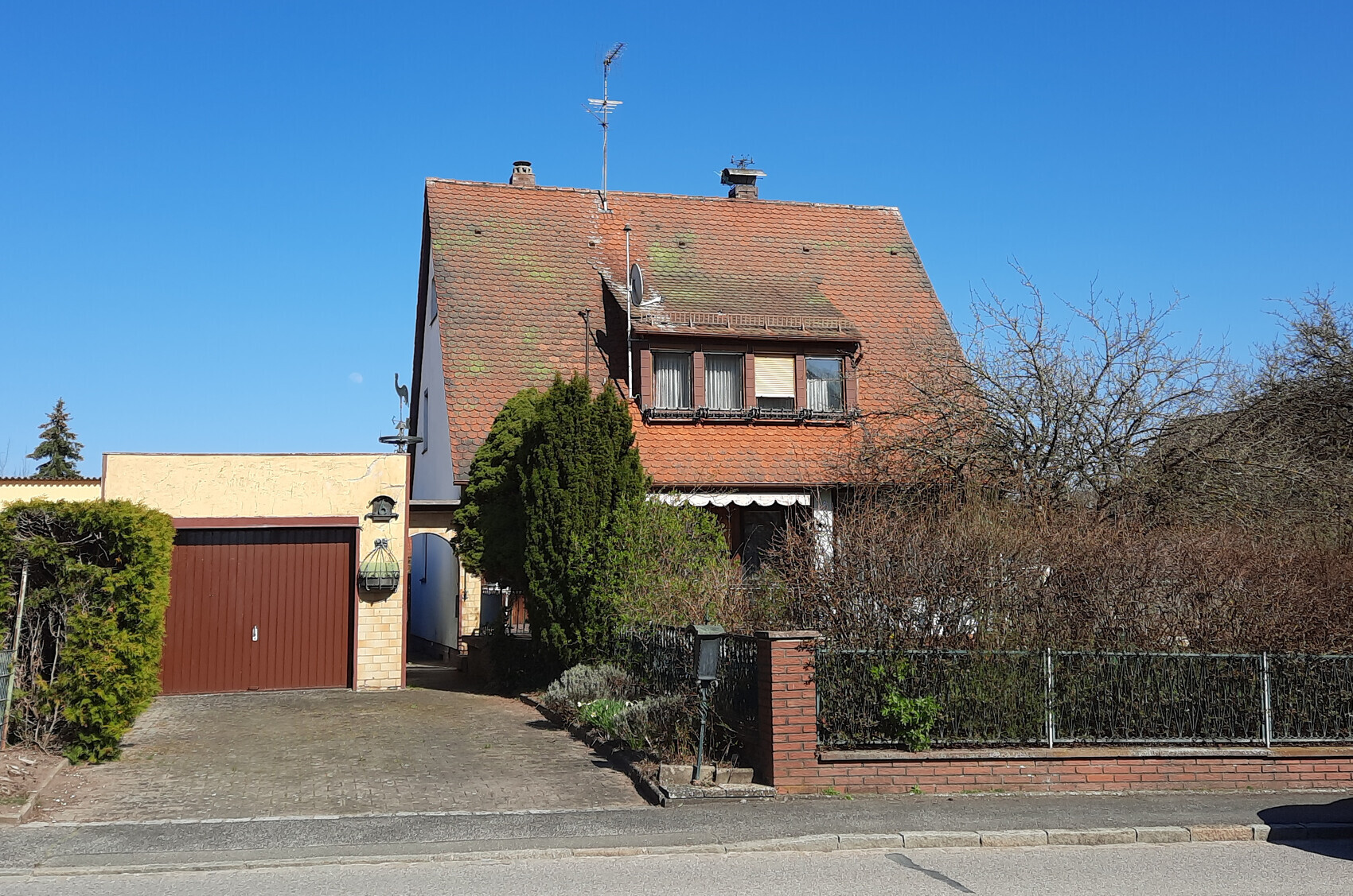 Zweistöckiges Haus mit rotem Ziegeldach, Garage links, Hecke und kahle Sträucher, blauer Himmel.