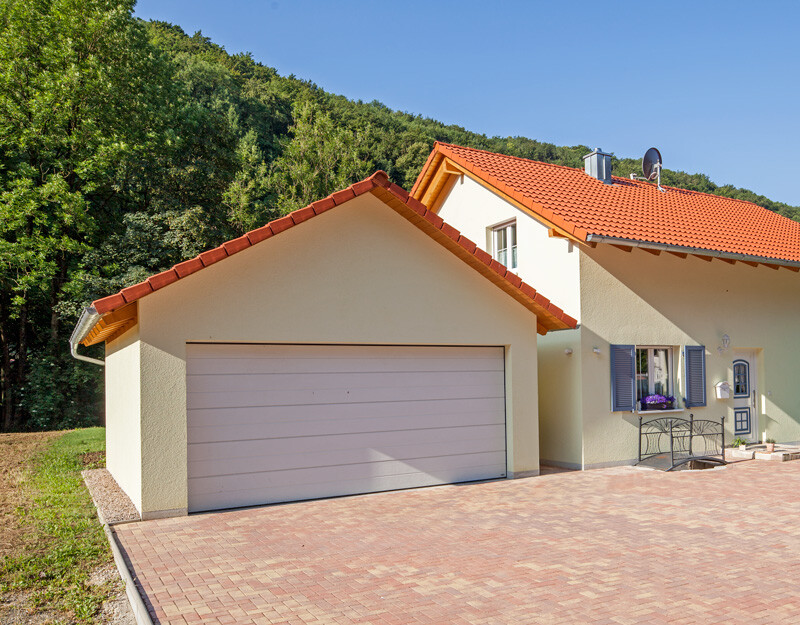Einfamilienhaus mit rotem Ziegeldach, Doppelgarage, gepflasterter Hof, blaue Fensterläden.