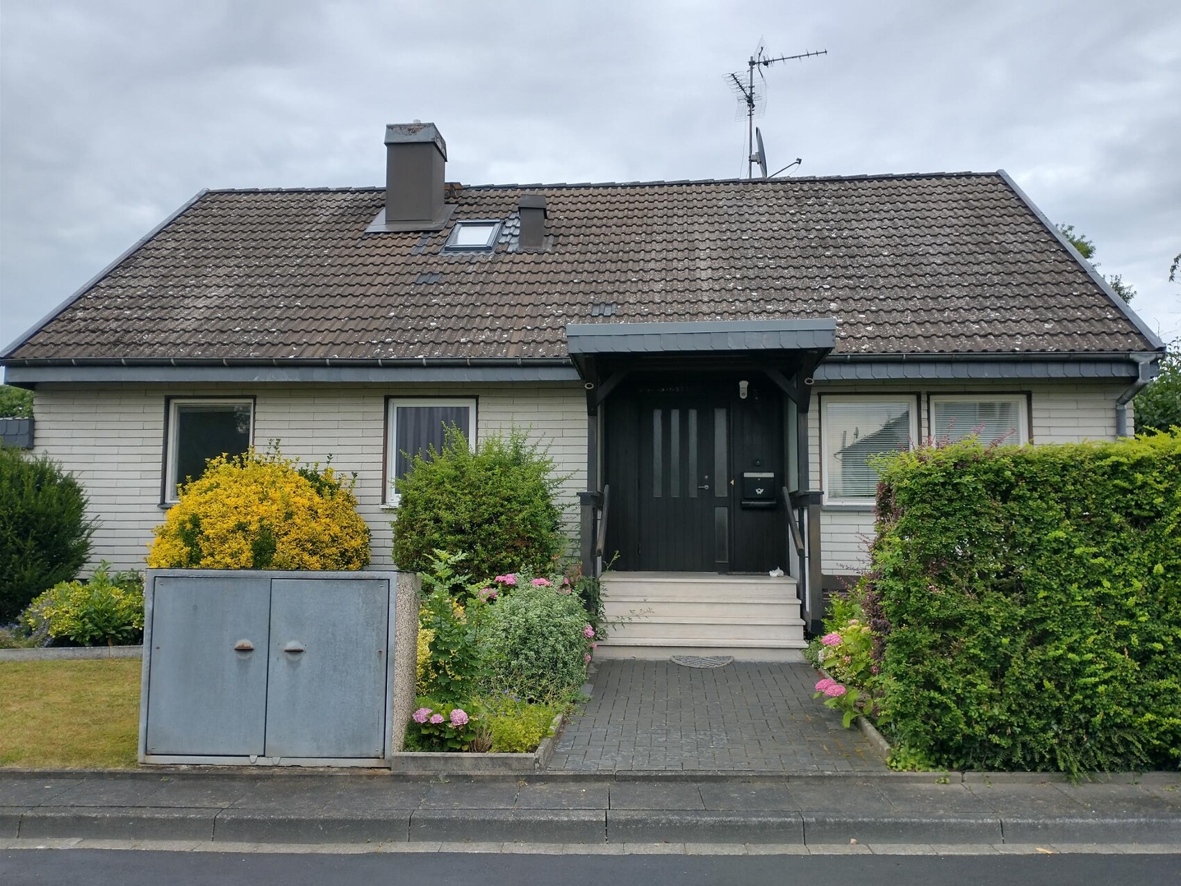 Vorderansicht eines Bungalows mit dunkler Tür, kleinem Vordach, Treppenstufen und Gartenhecke.