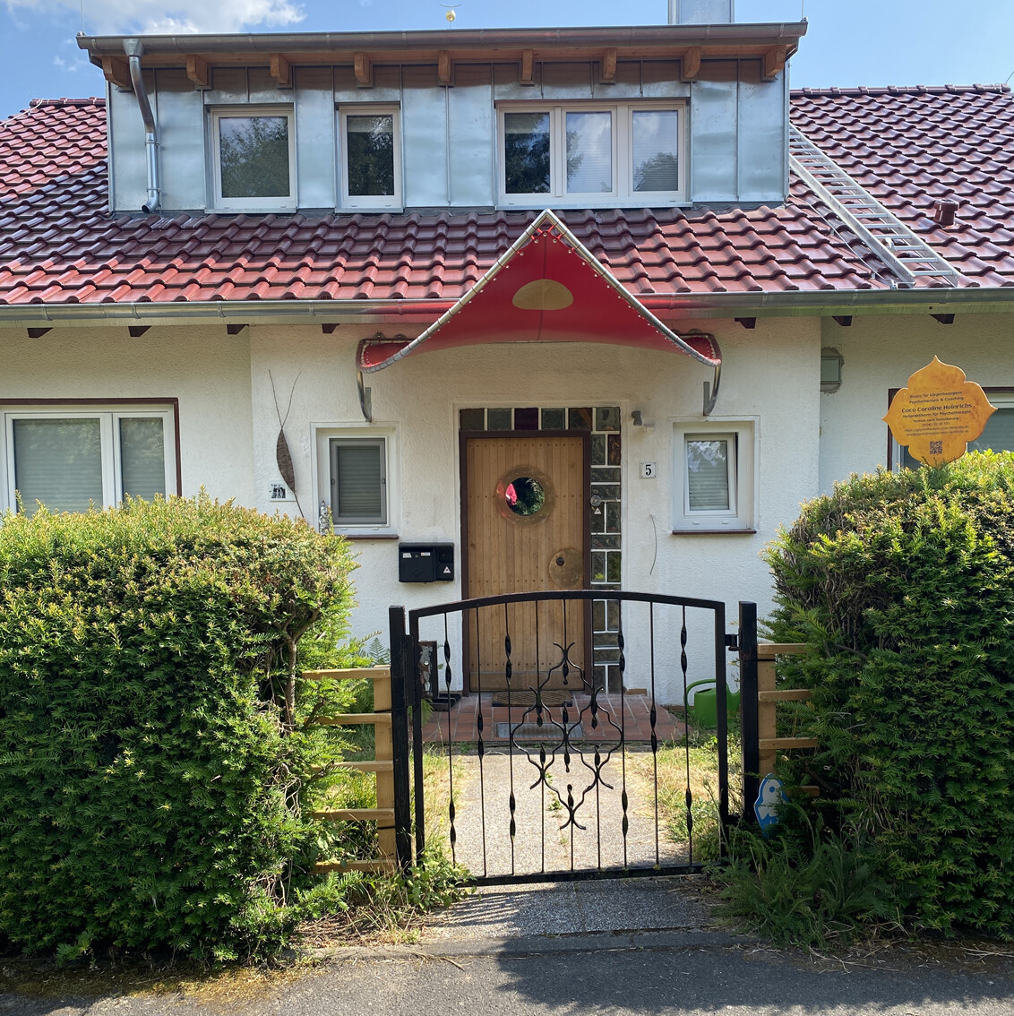 Hausfront mit rotem Segel-Vordach über Holztür, Fenstern, Gartenzaun und Hecken.