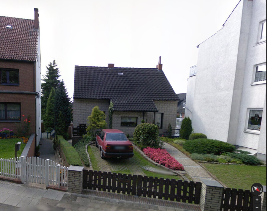 Rotes Auto parkt auf gepflastertem Hof vor kleinem Backsteinhaus mit Garten und pinken Blumen.