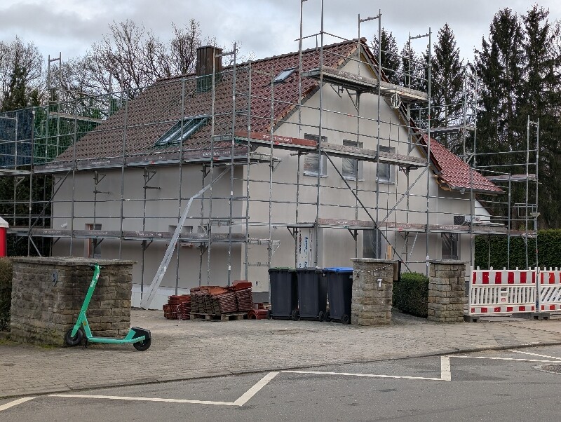 Ein Haus unter Renovierung mit Gerüst, Dachziegeln, Mülltonnen und grünem Tretroller davor.