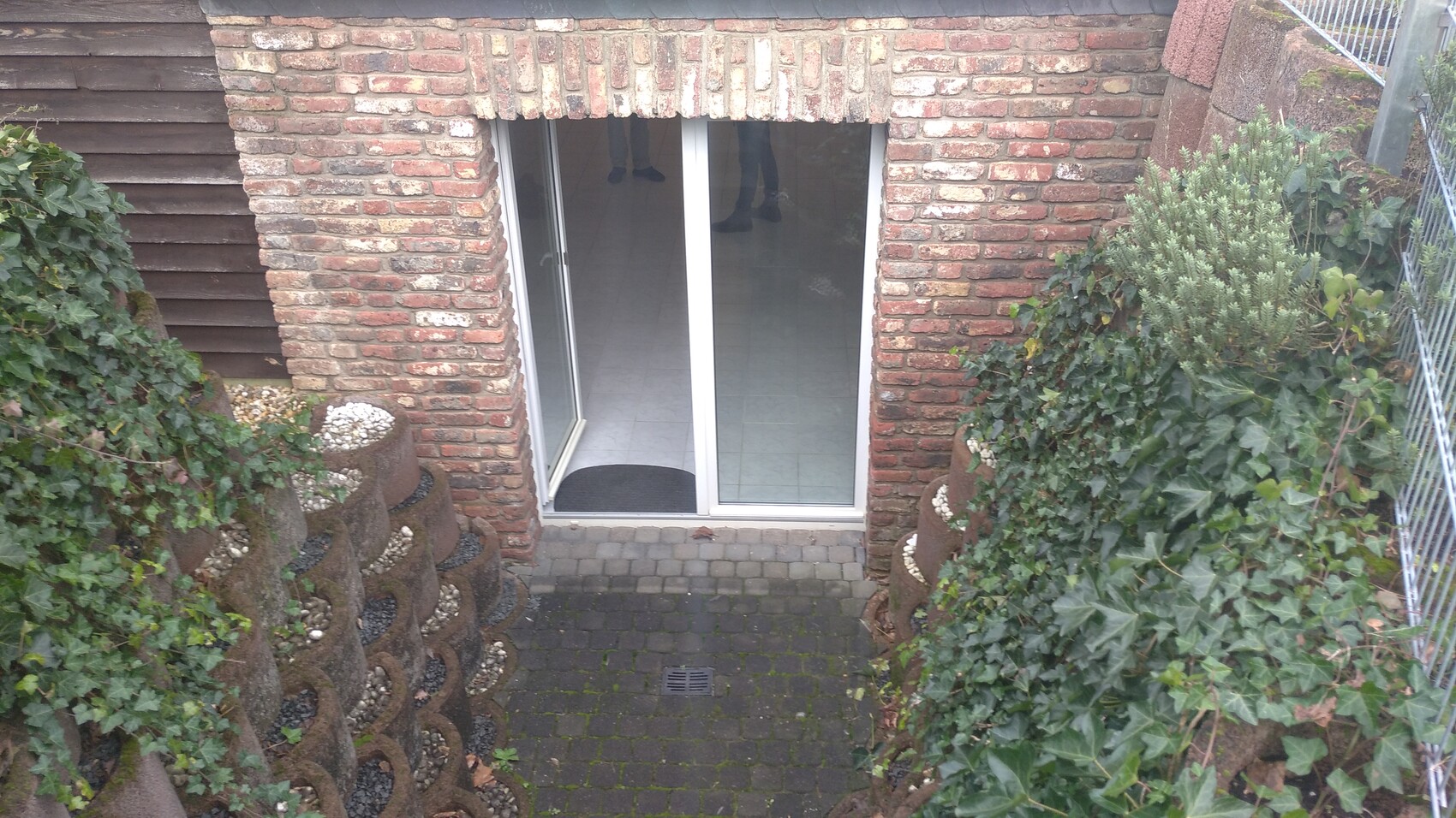 Backsteinfassade mit Glas-Schiebetür, Efeu bepflanzte Kübel beidseitig, gepflasterter Vorhof.