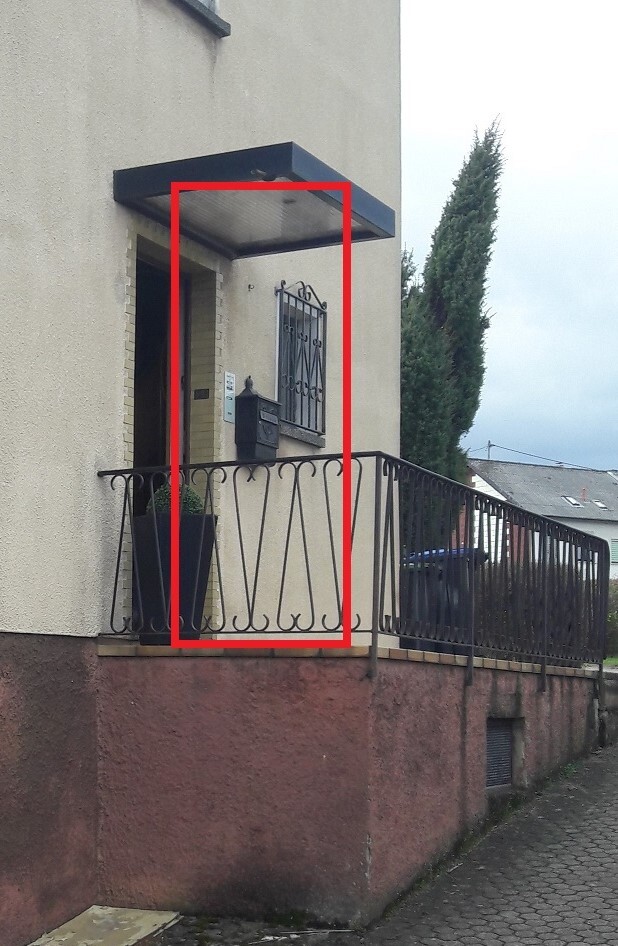 Eingang eines Hauses mit Überdachung, Tür, Fenster mit Gitter, Briefkasten und Geländer.