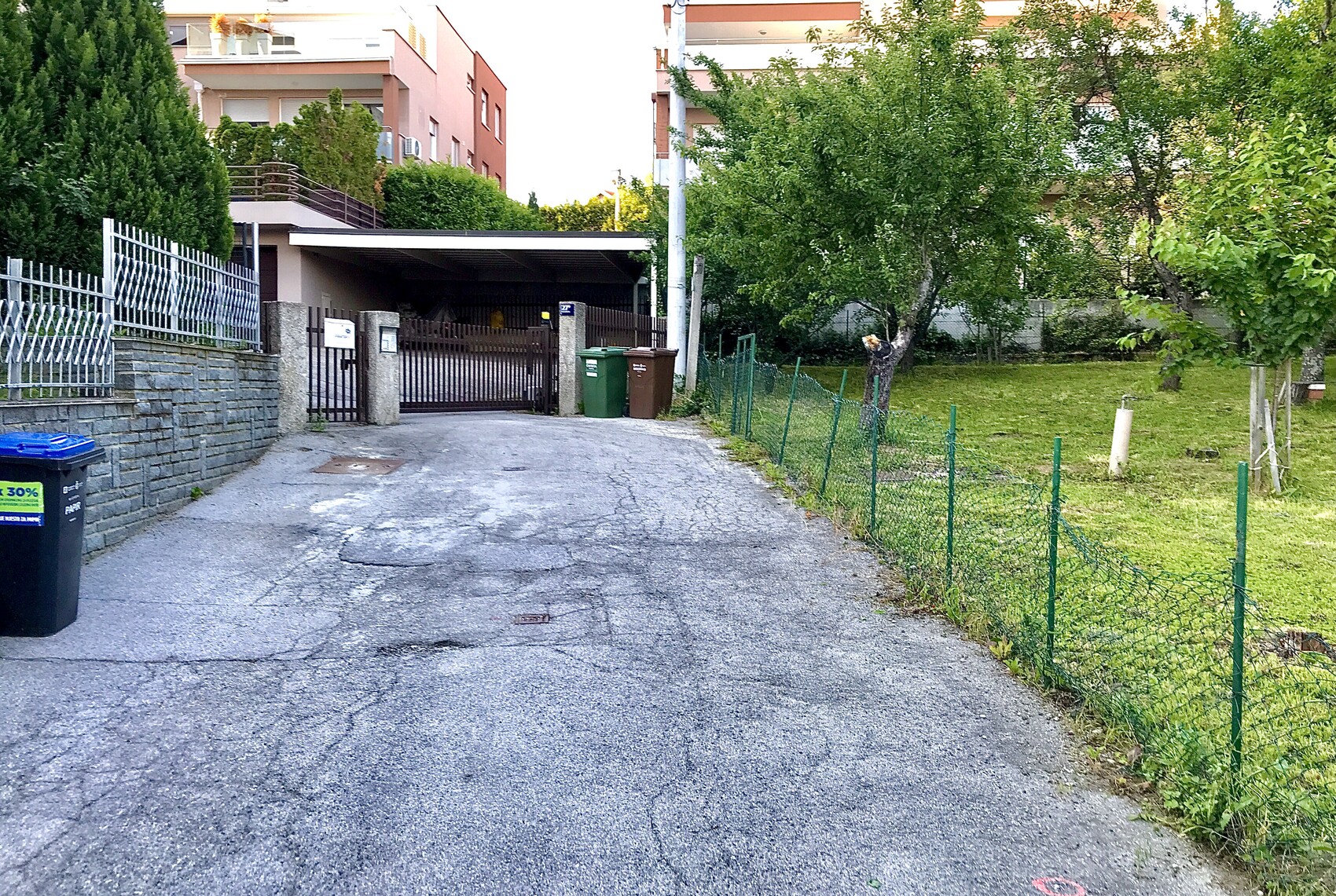 Zufahrt mit Tor zum Grundstück, rechts Garten mit Bäumen, links Mauer und Mülltonnen.