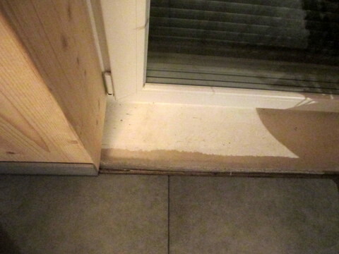 Türschwelle vor grauen Bodenfliesen, links Holzfront, daneben eine Glasfront mit Rollladen.