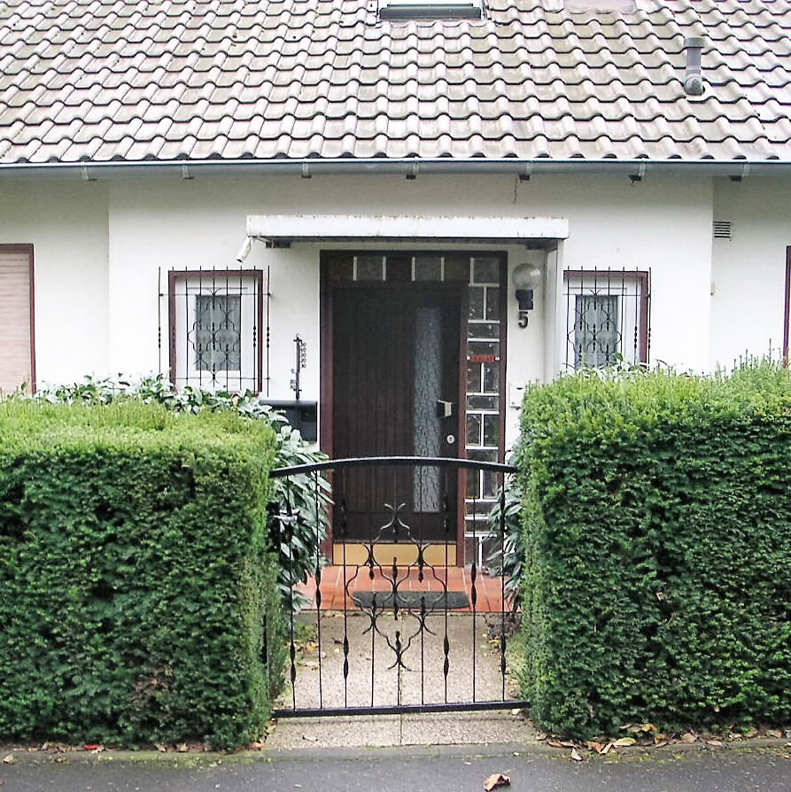 Vorderansicht eines Hauses mit Eingangstür, Tor und von Hecken gesäumtem Weg.
