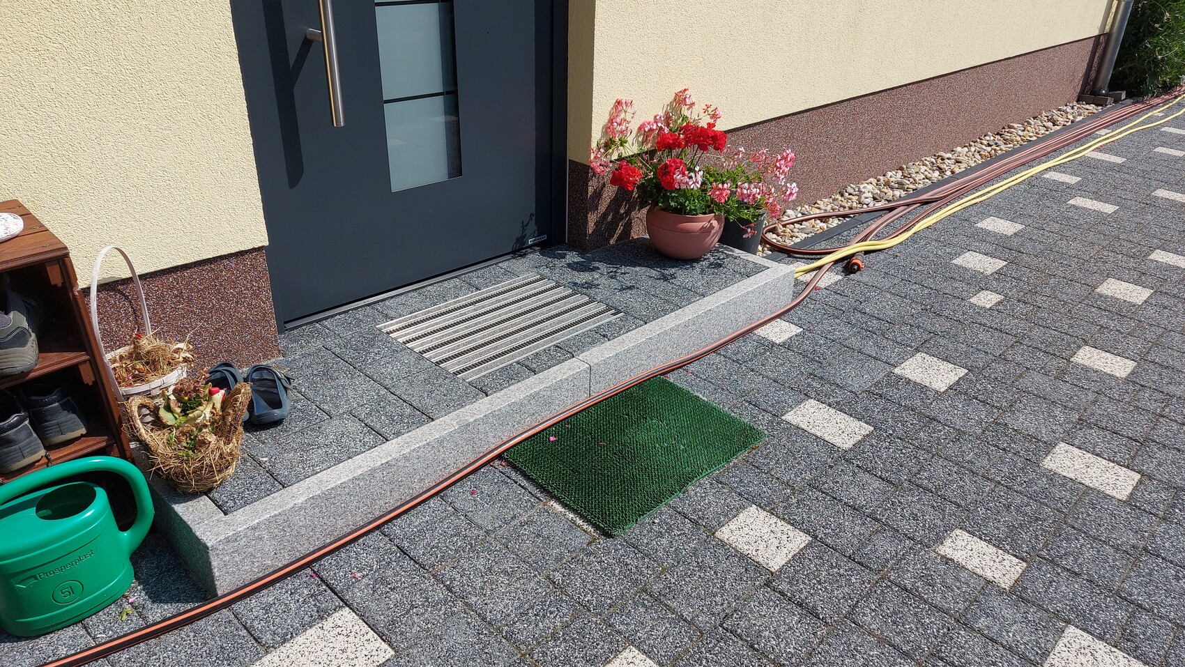 Vorderansicht eines Hauses: dunkle Tür, Blumenkübel mit roten Blüten, Schlauch und Schuhregal.