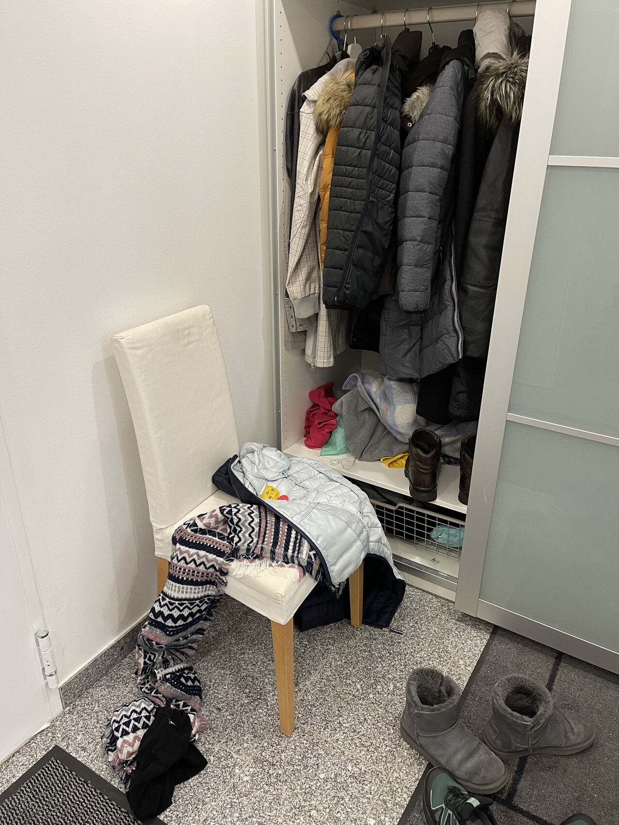 Offener Kleiderschrank mit Jacken und Mänteln; davor Stuhl mit Decke und Kleidung, Schuhe.