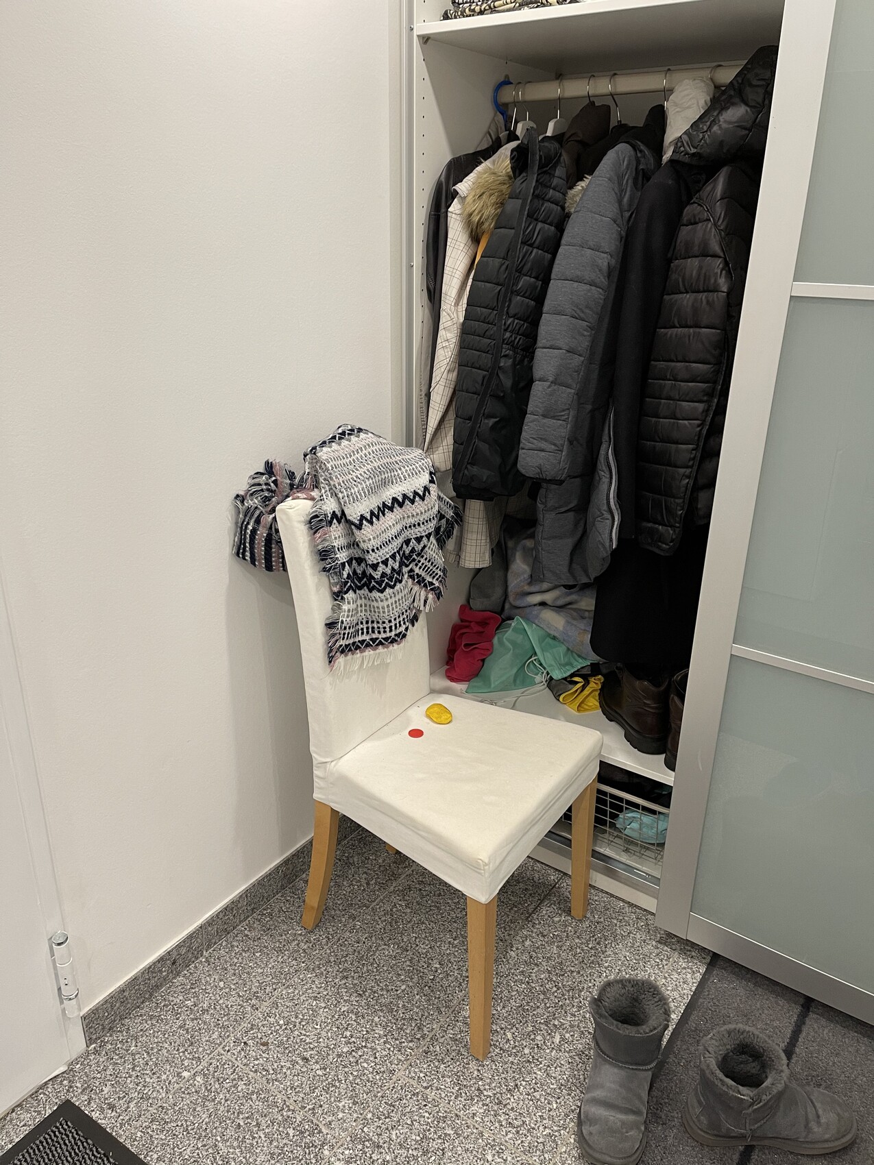 Offener Kleiderschrank mit Mänteln und Jacken; davor weißer Stuhl mit Schal, Schuhe daneben.