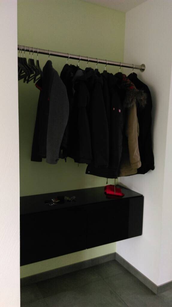 Garderobe mit Mänteln an einer Stange; darunter schwarze Schränke, rote Tasche darauf.