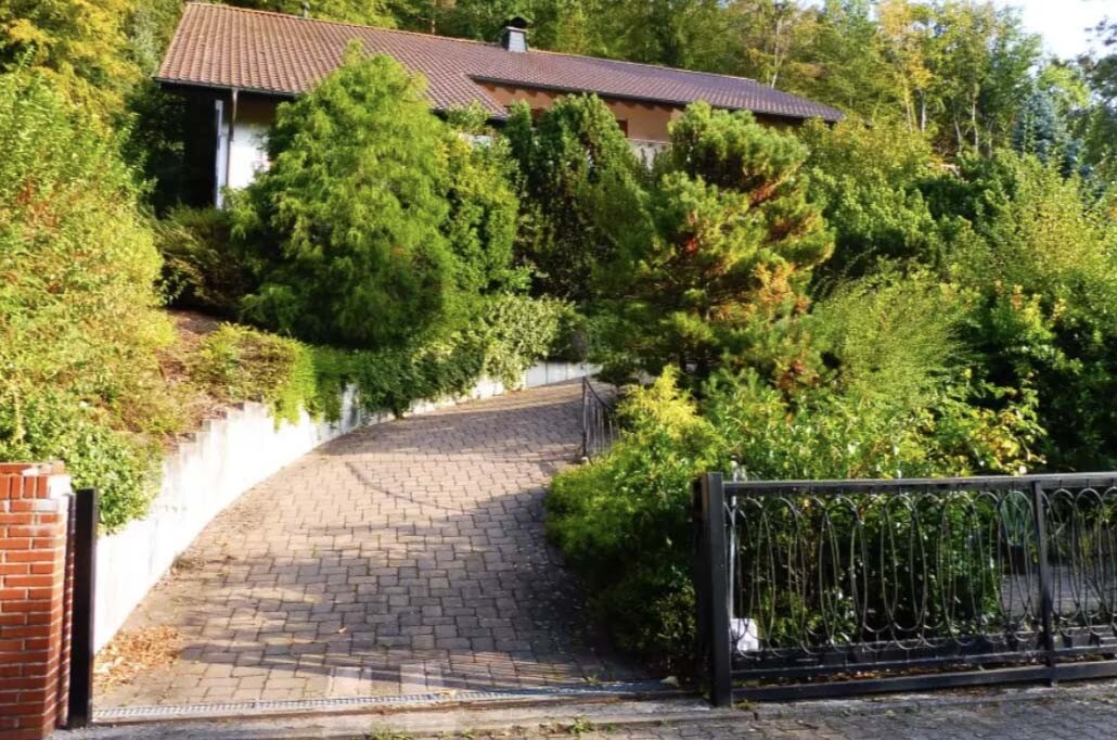 Gepflasterter Weg führt zu einem Haus, von dichtem Grün und einem Metallzaun umgeben.