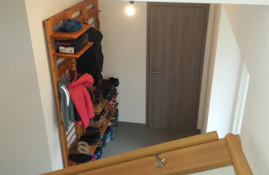 Kleiner Eingangsbereich mit Holzschuhregal, Mantel, Schuhen und Tür im Hintergrund.
