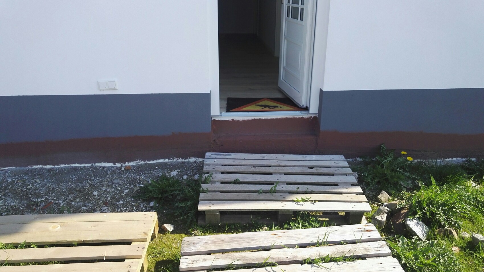 Offene Haustür eines weißen Hauses; gelbes Warnschild auf der Fußmatte, Palettenweg zum Eingang.