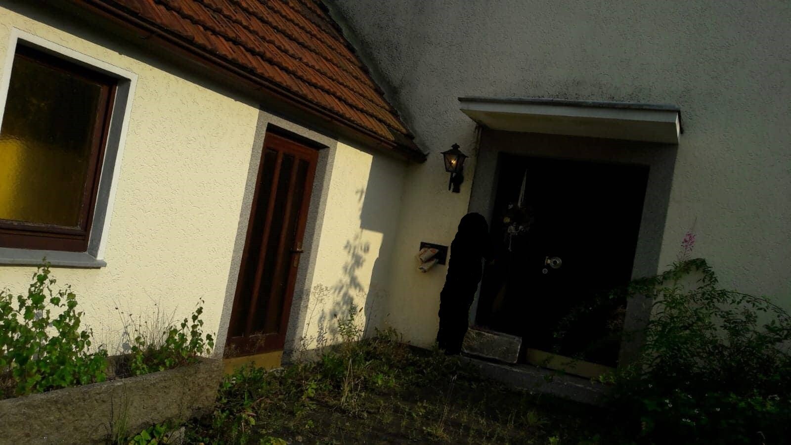 Person in schwarzer Kleidung steht vor dunkler Eingangstür eines Hauses; Fenster links.