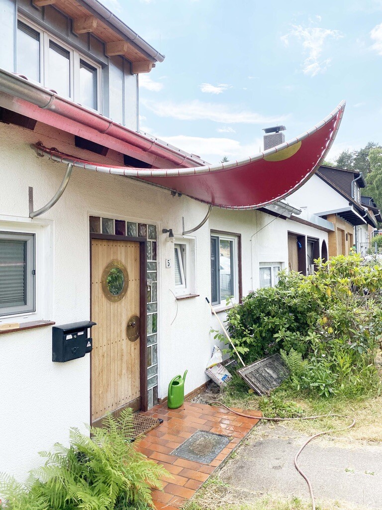 Vorderansicht eines Hauses mit Holztür, rotem Bogendach über dem Eingang und Gartenpflanzen.