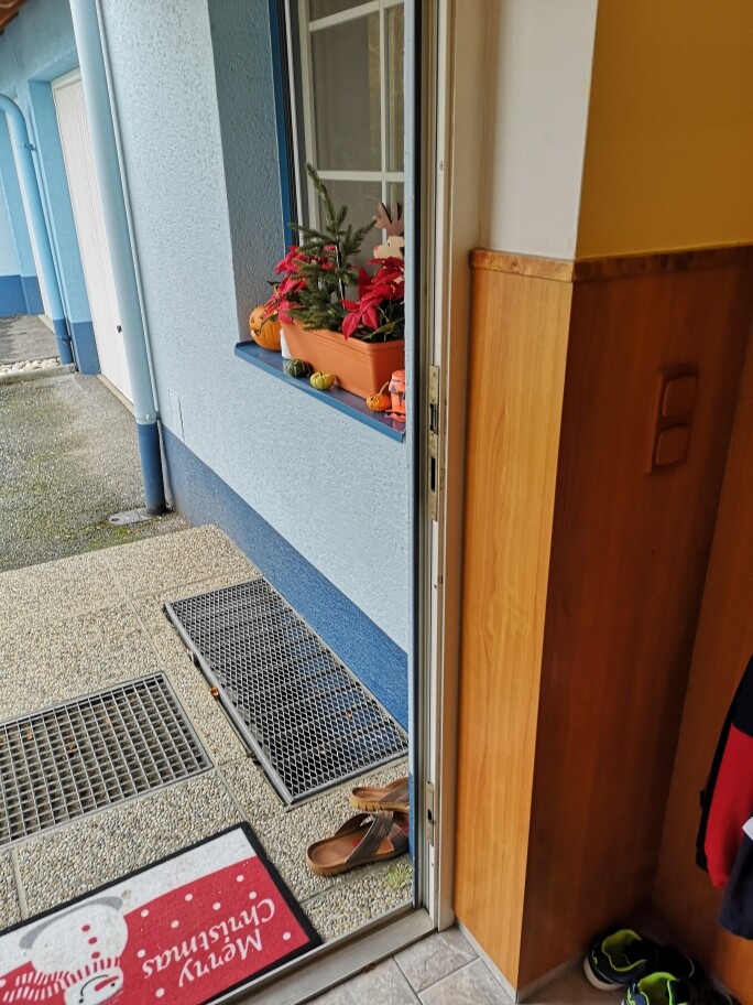 Offene Tür zu blauem Hausflur; Fensterbank mit roten Blumen und Kürbissen, Sandalen davor.