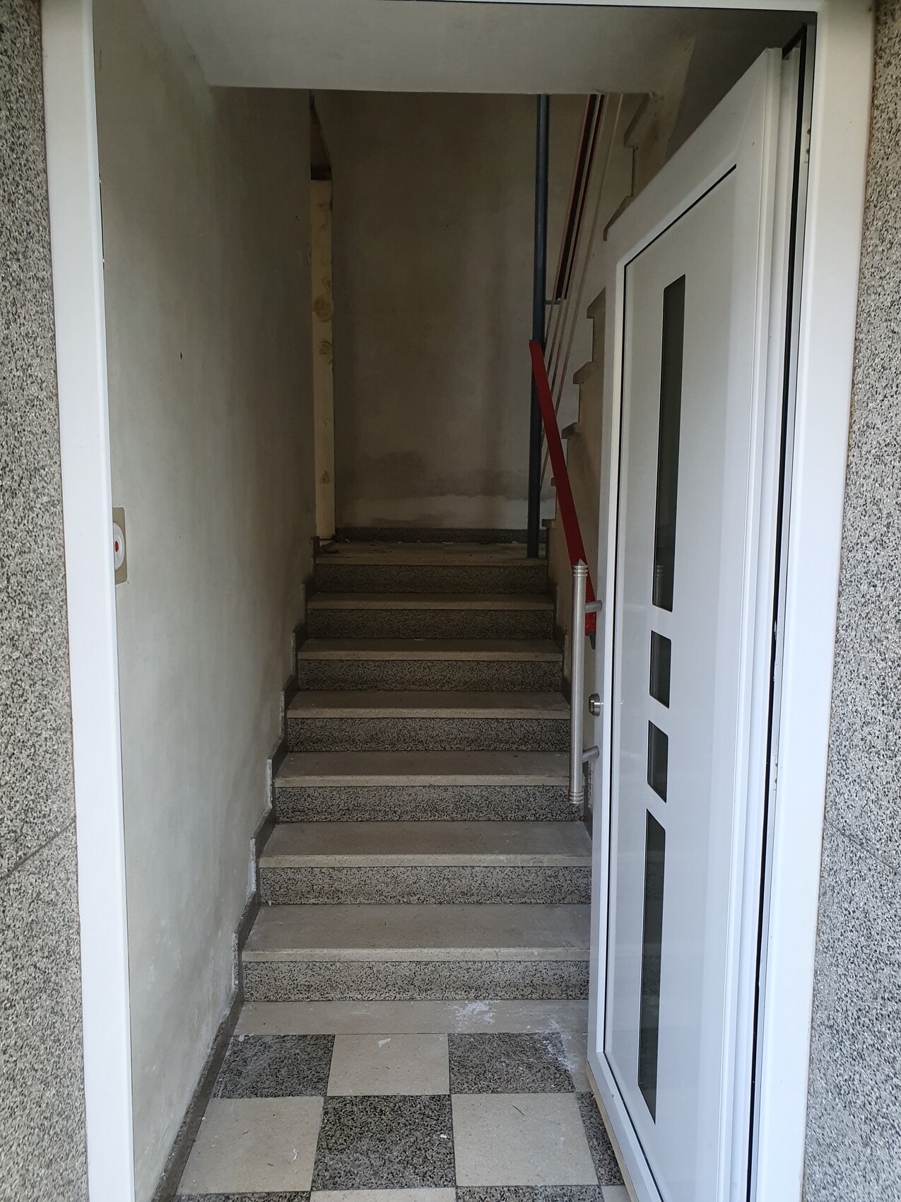 Heller Eingangsbereich mit Treppe nach oben, grauen Stufen, rotem Geländer rechts und offener Tür.